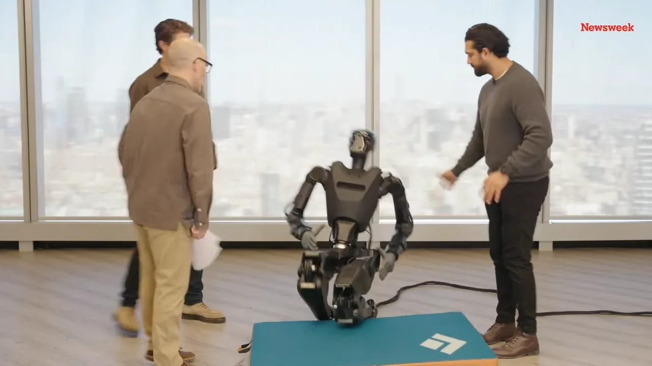 A screenshot from a video demonstration showing the Phantom robot collapsing toward the floor. Standing nearby and watching the fall are Foundation co-founder Mike LeBlanc, CEO Sankaet Pathak, and a Newsweek reporter. The setting appears to be an office environment.