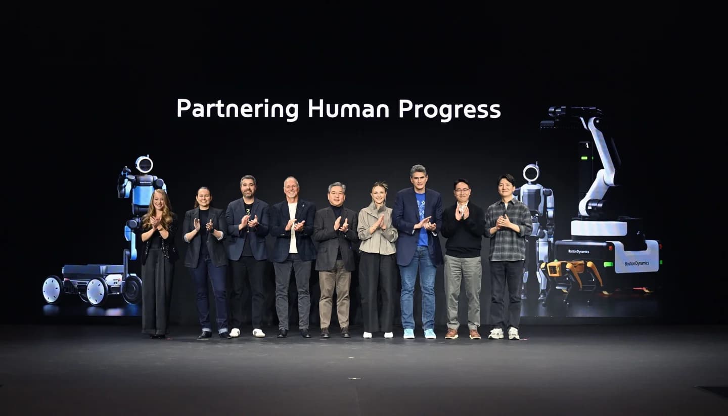 A group of nine executives and engineers standing on a stage beneath the digital banner "Partnering Human Progress". Digital renderings of the Atlas, Spot, and Stretch robots are visible on the screen behind them.