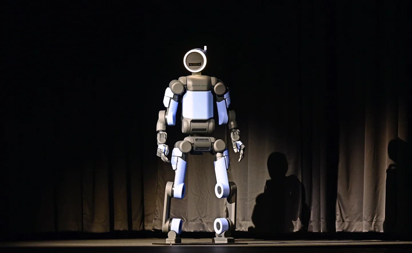 A full-length shot of the all-electric Boston Dynamics Atlas humanoid robot standing centered on a dark stage. The robot has a blue and black chassis and a circular head illuminated by a bright white ring light.