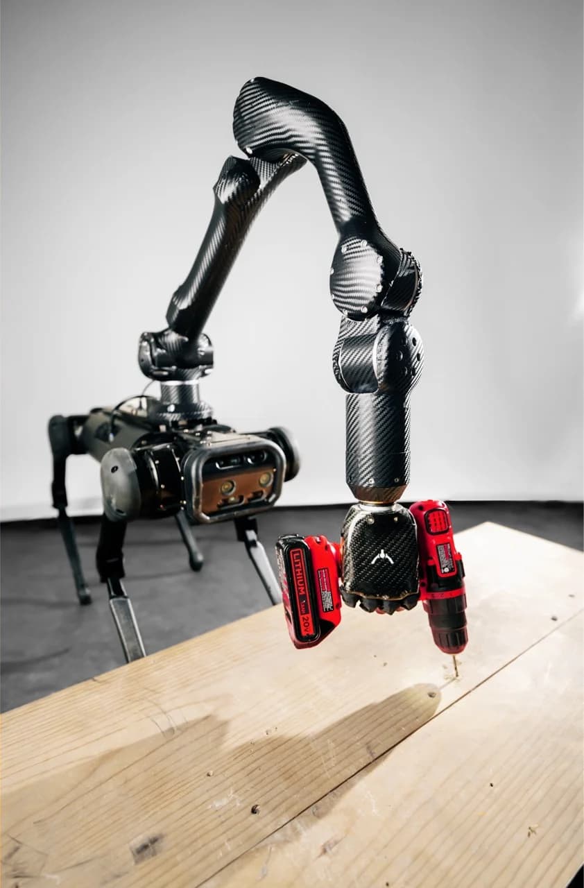 A carbon-fiber robotic arm attached to a mobile robot dog uses an Ability Hand to firmly grip and operate a red power drill against a wooden plank.