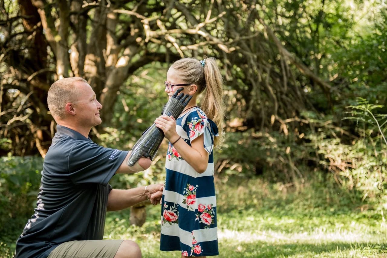 A man wearing a carbon-fiber PSYONIC Ability Hand prosthetic kneels outdoors, gently touching the face of a young girl who is smiling.