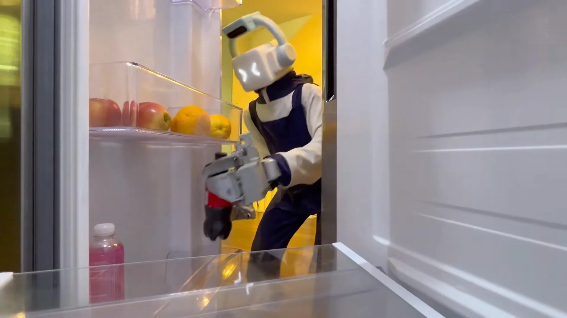 A humanoid robot wearing a blue apron and equipped with an articulated robotic hand reaches inside an open refrigerator to retrieve a bottle from a door shelf.