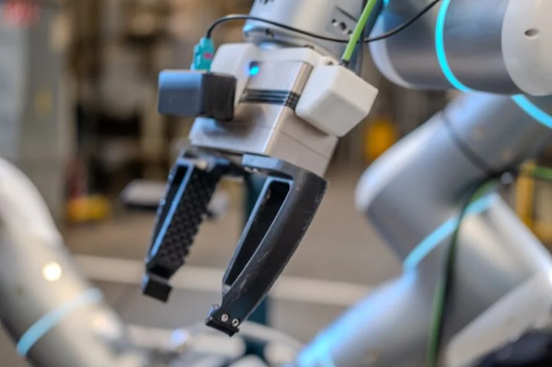 A close-up of a two-fingered robotic gripper with a black camera module and white sensors mounted on the wrist assembly.