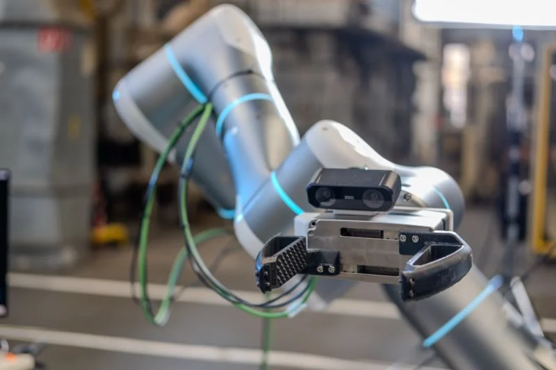 Side view of a silver robotic arm with blue LED rings, featuring a ZED stereo camera mounted directly above a black parallel gripper.
