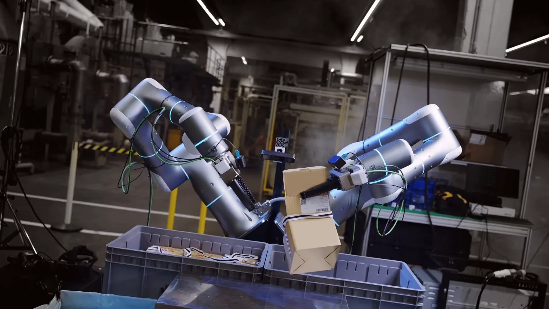 A bimanual robotic system with two silver arms lifting a cardboard box in a dimly lit factory workstation.