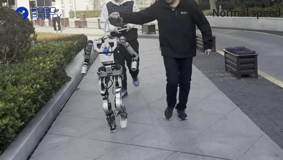 A bipedal humanoid robot prototype with an exposed mechanical frame runs on an outdoor walkway while being monitored by two researchers using a safety tether.