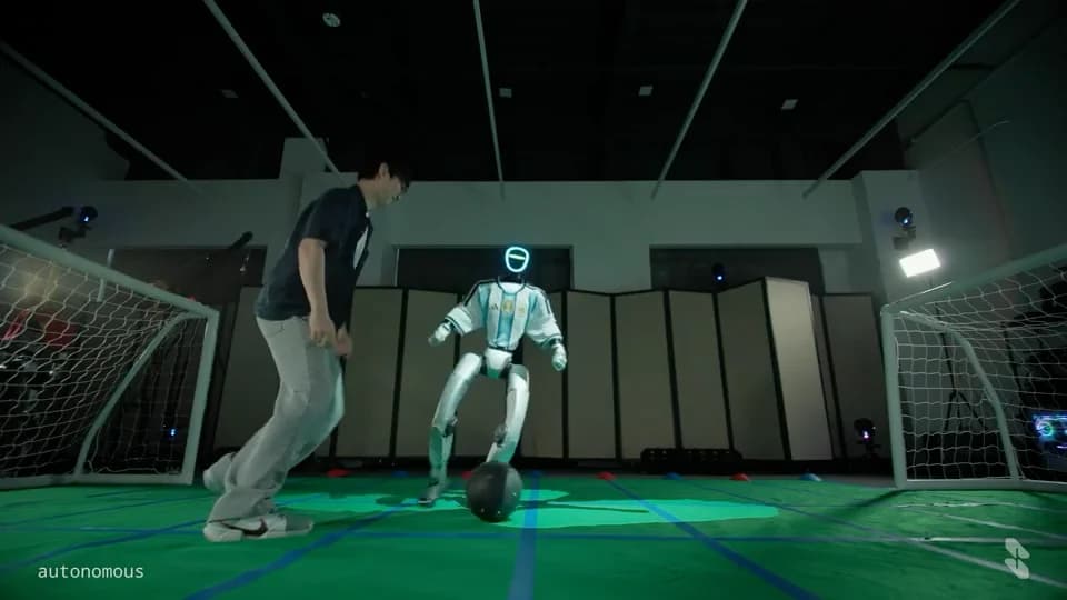 Unitree G1 humanoid in a blue-and-white soccer jersey facing a person, with a ball at its feet between two indoor goals.