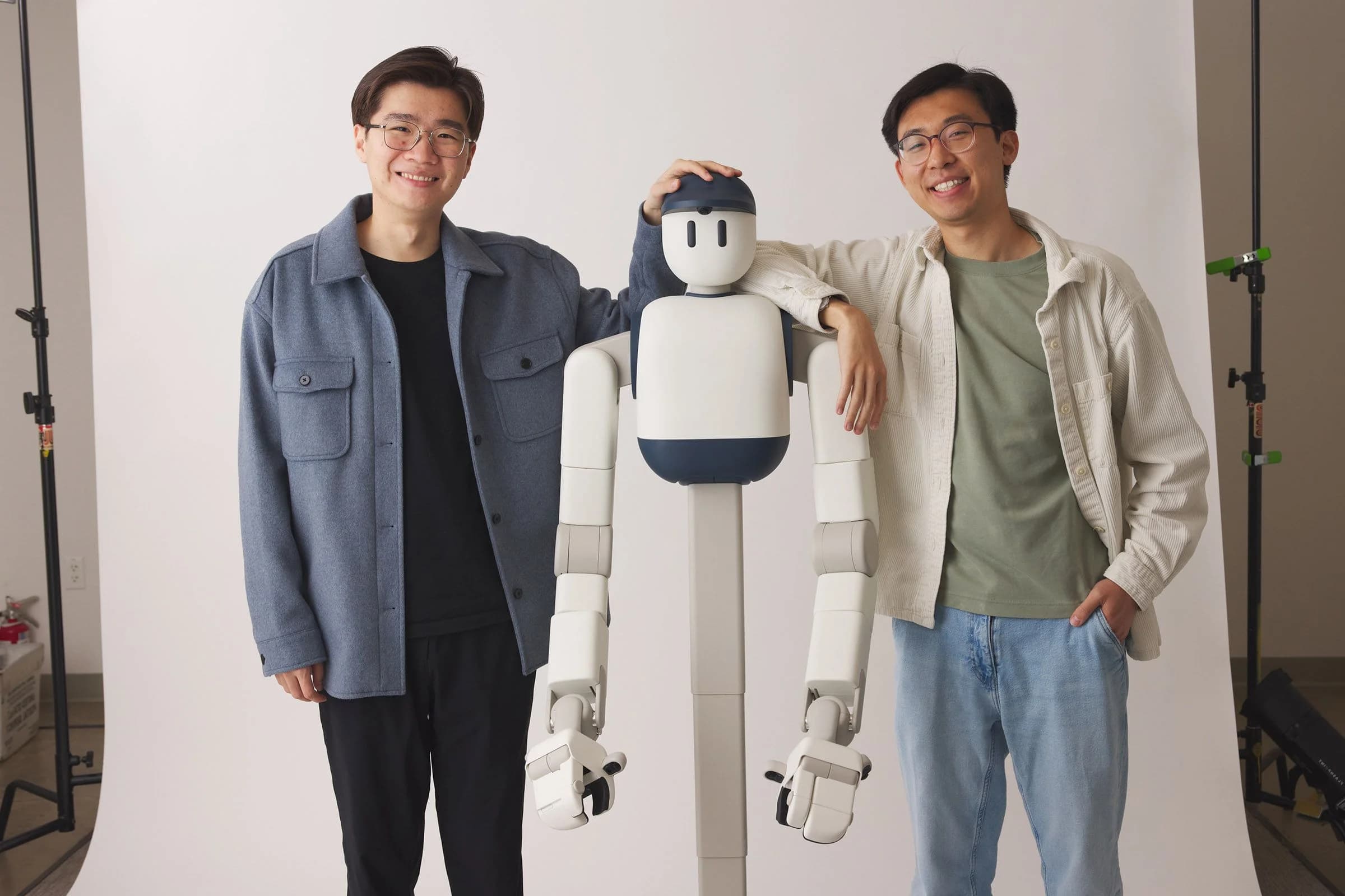 Sunday Robotics co-founders Tony Zhao (left) and Cheng Chi (right) smiling and standing next to their white and blue domestic robot, Memo, which features a wheeled base and telescoping spine.