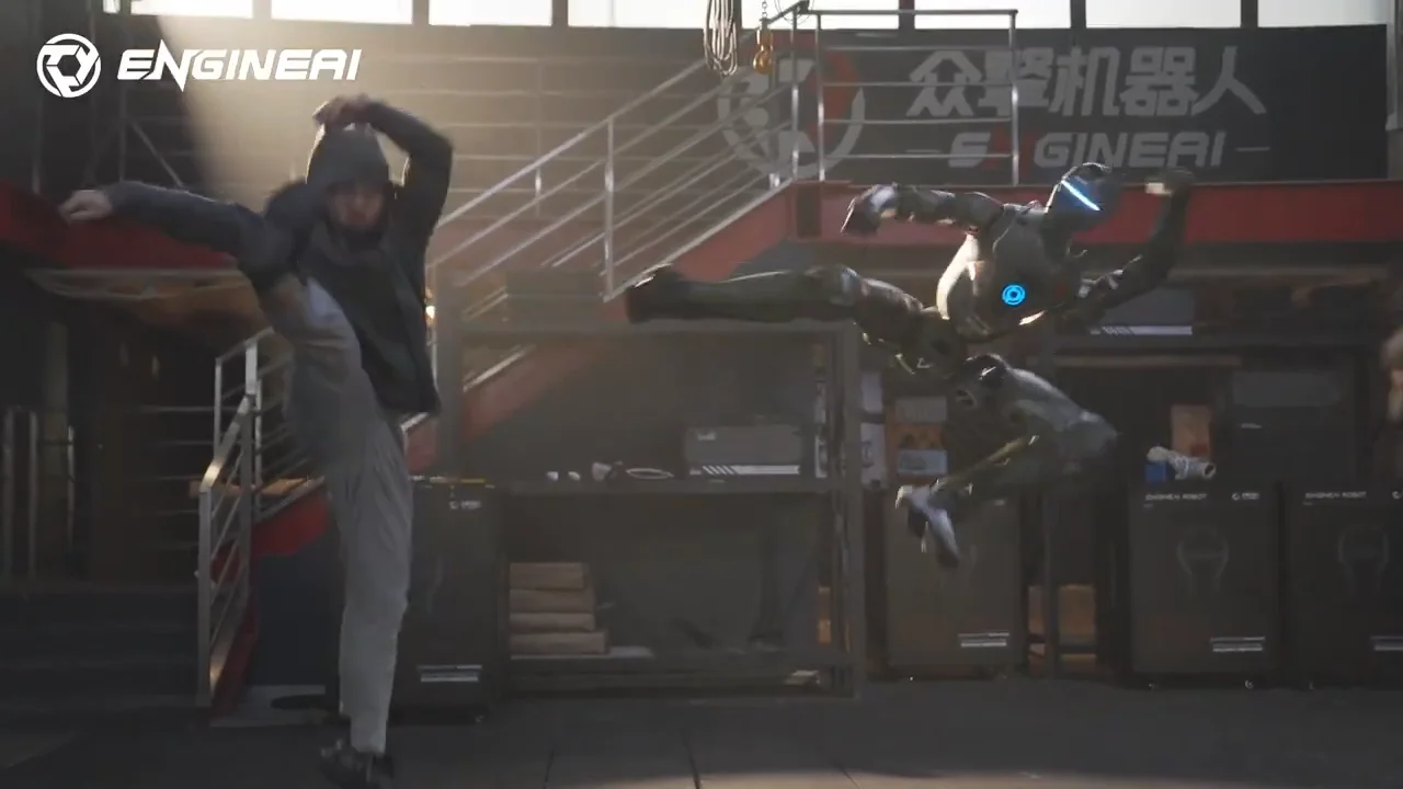 A behind-the-scenes shot showing a human martial artist and the EngineAI T800 robot performing synchronized flying side kicks in an industrial gym setting.