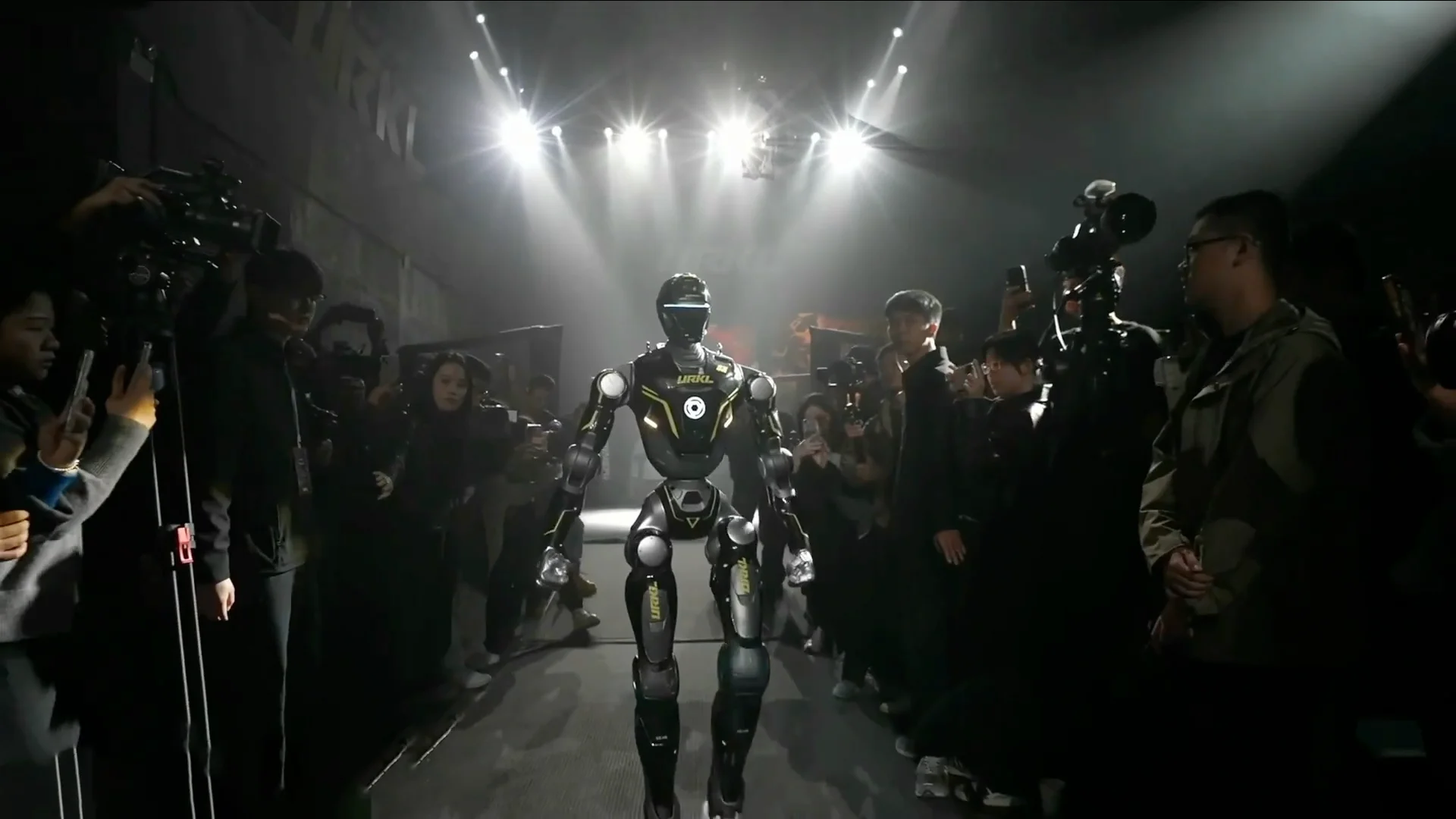 An EngineAI T800 humanoid robot with a black chassis and yellow accents walks toward the arena stage at the URKL debut event, surrounded by media and spectators.