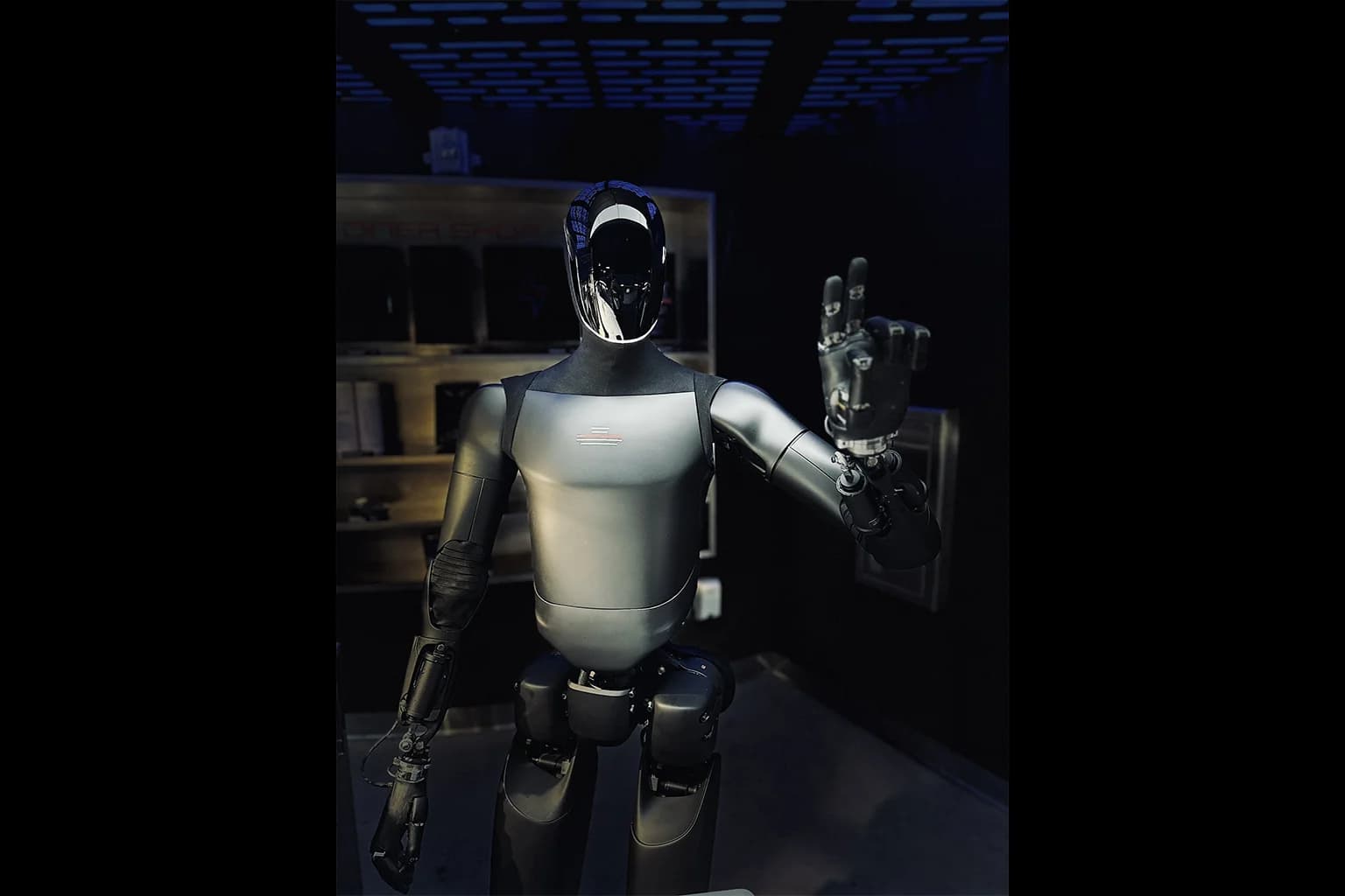 Front view of a Tesla Optimus Gen 2 humanoid robot standing inside a dimly lit interior space with its left hand raised in a greeting gesture, displaying a metallic silver torso and black articulated limbs.
