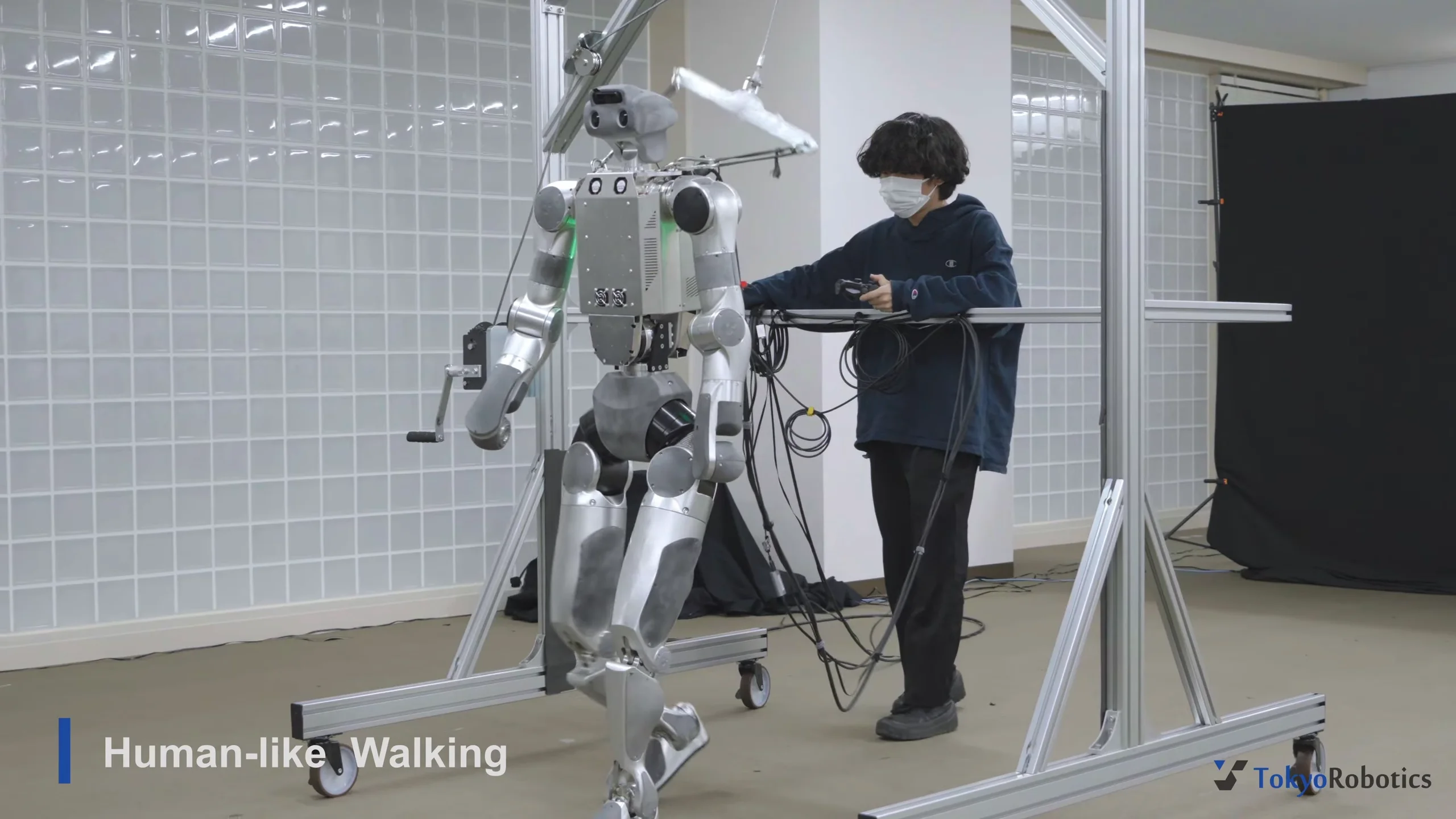 A silver bipedal humanoid robot walking within a metallic support frame while a researcher in a dark hoodie and face mask monitors the demonstration.