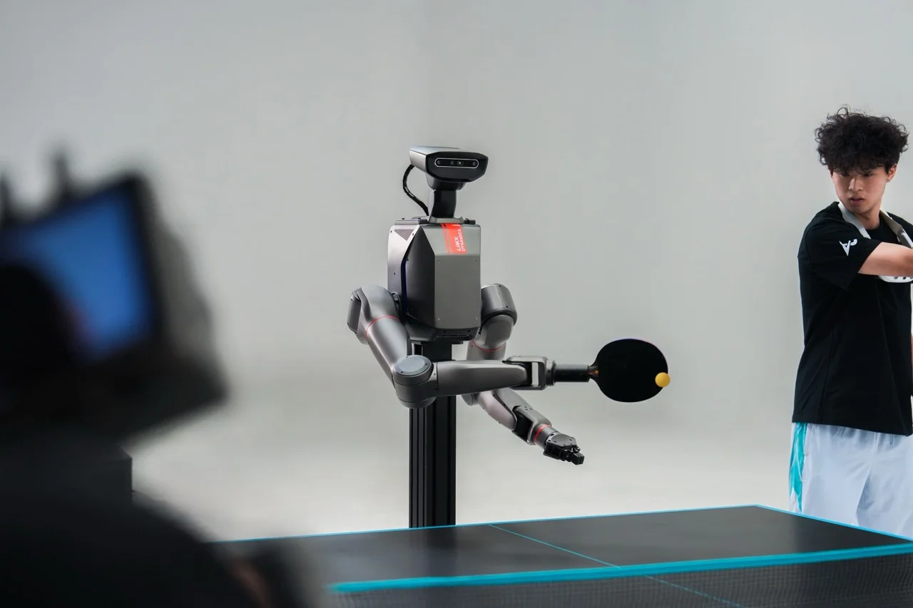 The TRON 2 robot in its stationary dual-arm configuration, mounted on a black pillar, holding a paddle and striking a table tennis ball during a match against a human.