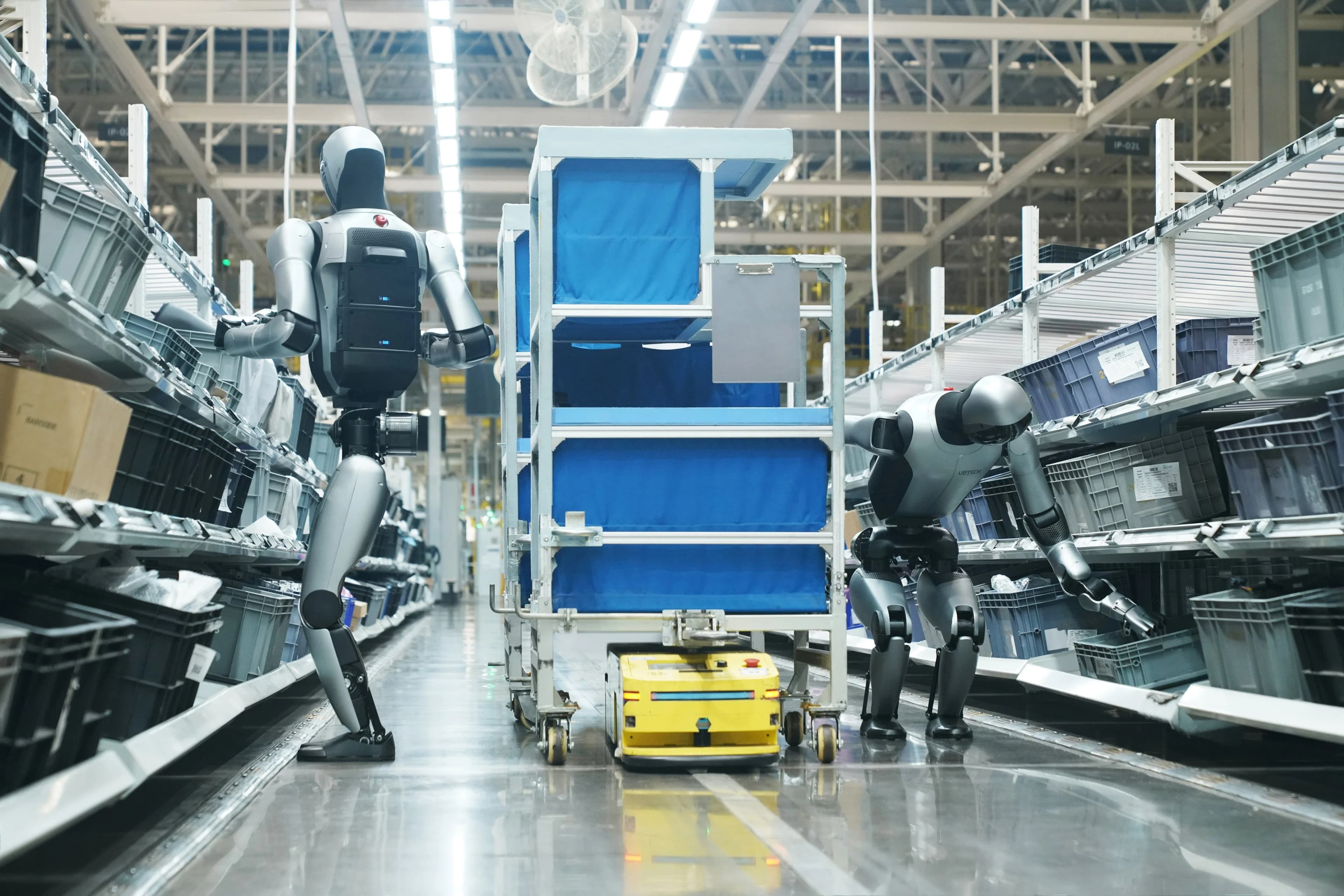 Two humanoid robots working in a narrow factory aisle alongside a small yellow autonomous mobile robot (AMR) positioned under a blue cart. The robot on the right bends down to interact with a lower shelf, while the robot on the left stands upright near the cart.