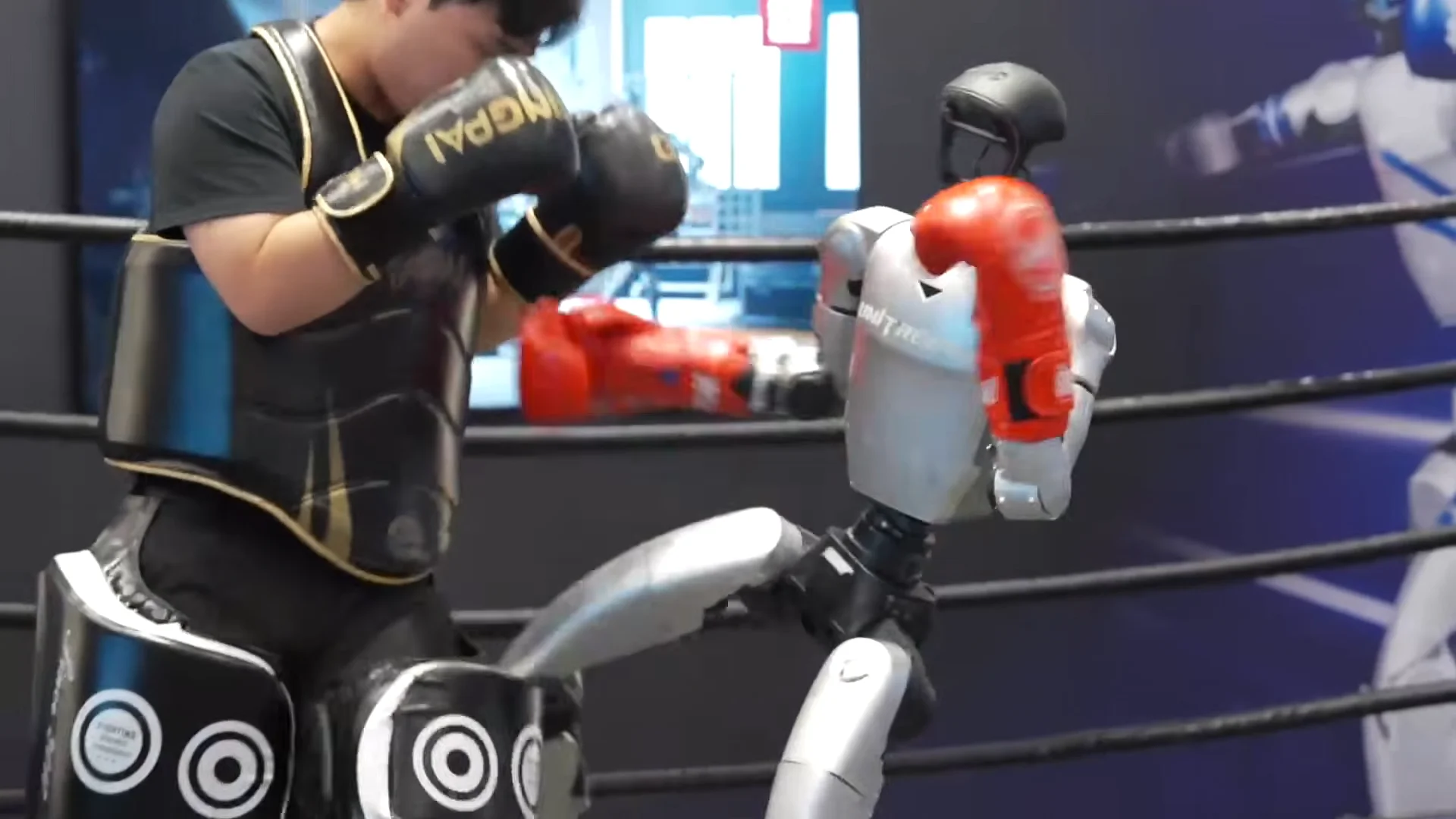 A silver Unitree G1 humanoid robot wearing red boxing gloves balances on one leg inside a ring while delivering a body kick and punch combination against a human trainer wearing protective black chest and thigh gear.