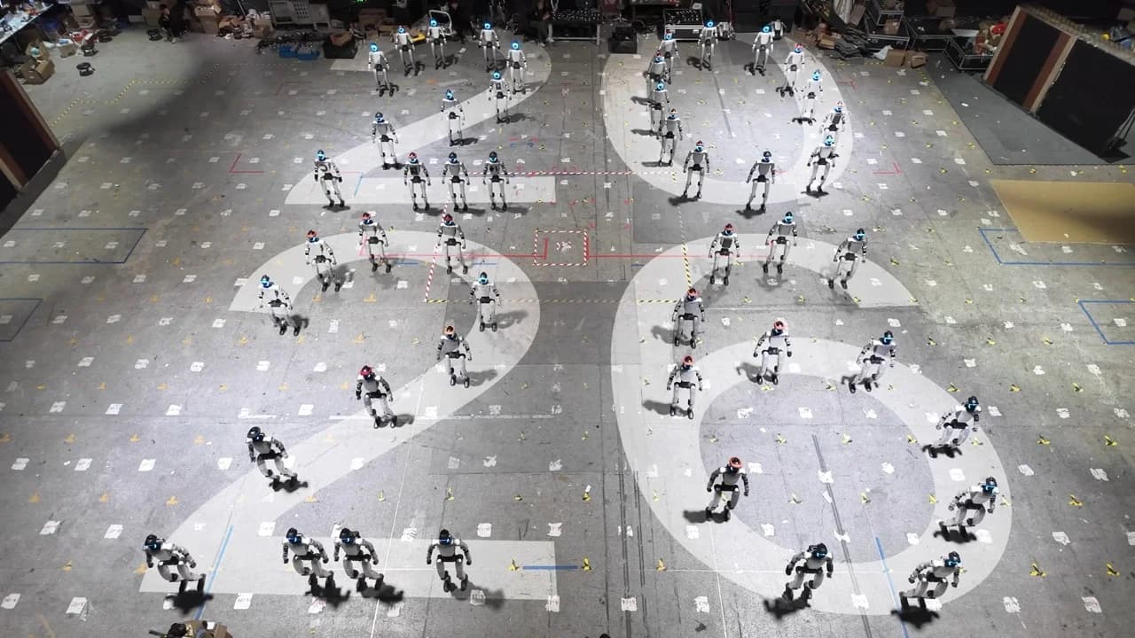 An overhead, top-down view of a large studio floor where dozens of silver G1 humanoid robots are precisely positioned to form the number "2026" amid various markings.