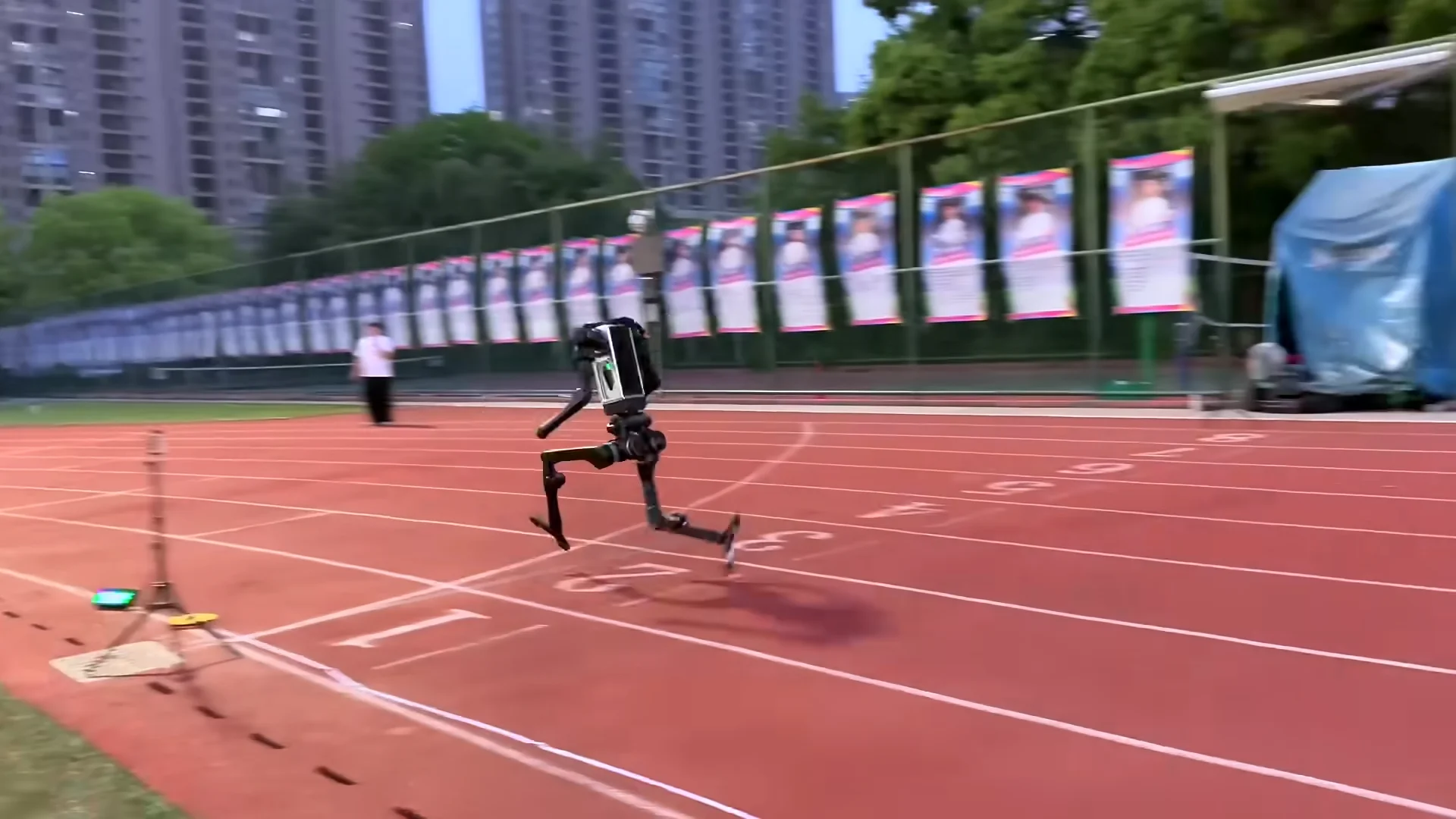A side view of a black humanoid robot, identified as the Unitree H1, sprinting on a red athletic track. The robot is captured mid-stride with both legs bent and its feet momentarily off the ground. In the background, a person in a white shirt stands on the track, and a row of banners is attached to a fence in front of a line of trees and tall apartment buildings.