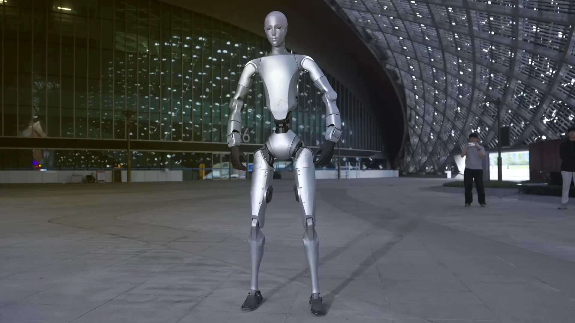 A full-body shot showing the silver Unitree H2 humanoid robot standing outdoors on a paved plaza. The robot features a narrow metallic waist, a stylized smooth silver head mask, and dark-colored basic end-effectors. In the background, a modern architectural building with glass and latticed facades is visible alongside two onlookers.