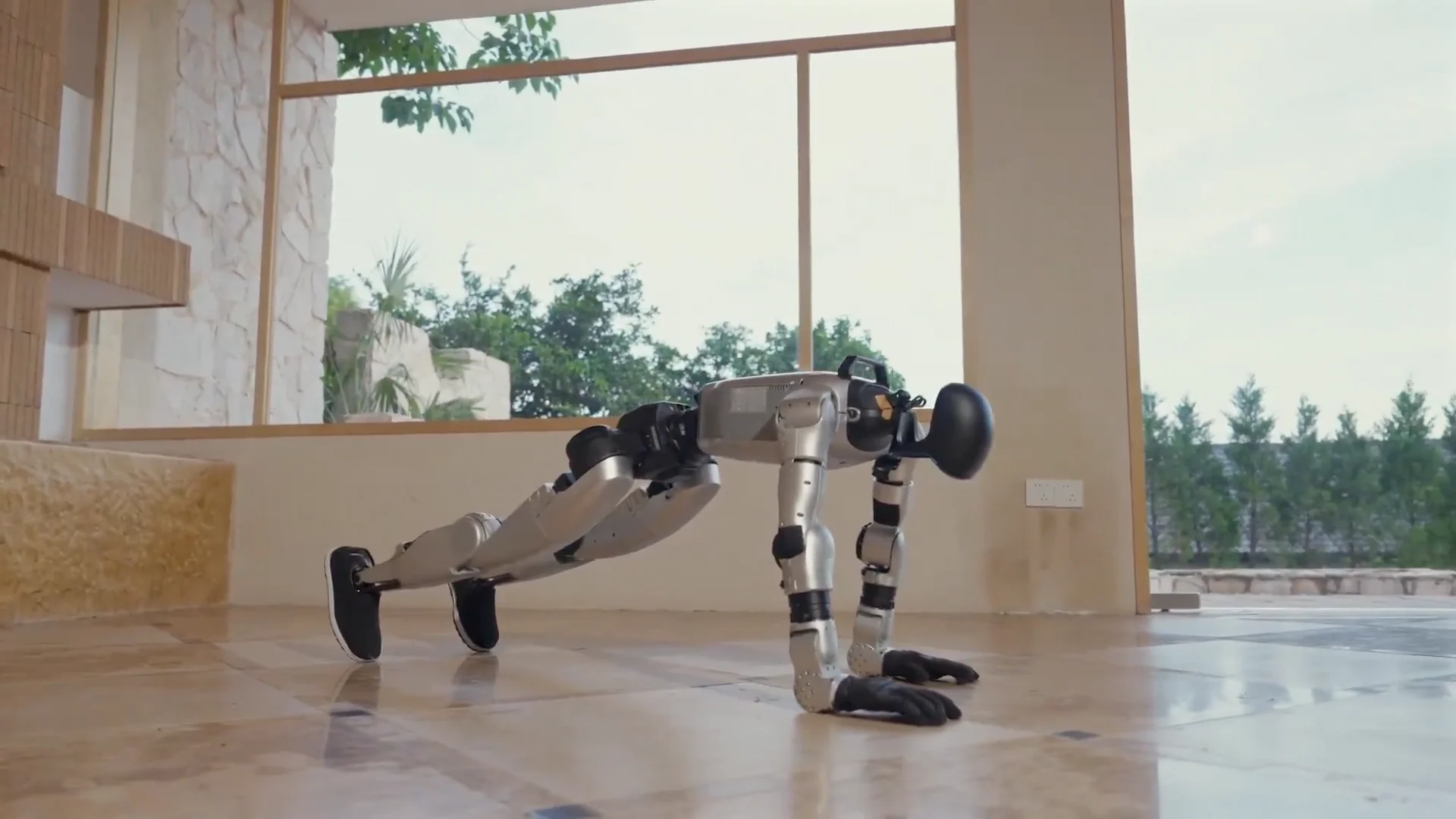 A silver and black Unitree G1 humanoid robot performing a push-up on a polished stone floor inside a modern room with large glass windows.