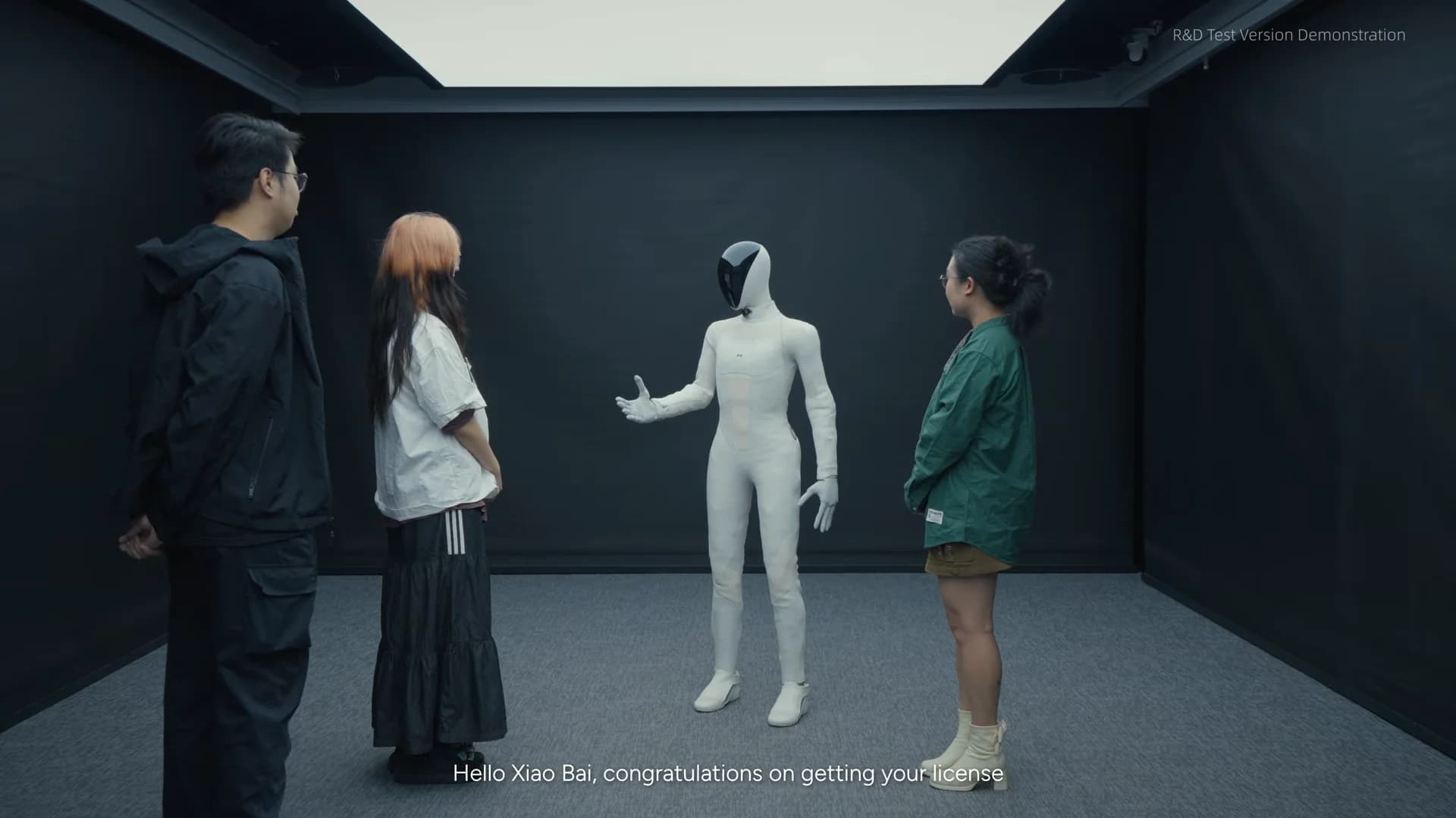 XPENG’s white IRON humanoid gestures toward three testers in a dark-walled lab during an interaction demonstration.