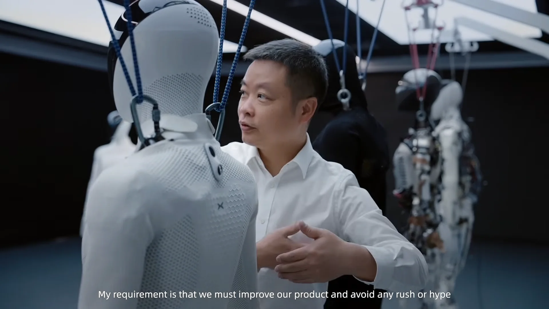 XPENG CEO He Xiaopeng speaks while gesturing near an IRON humanoid prototype suspended by overhead safety harnesses in an R&D lab. Subtitles read: 'My requirement is that we must improve our product and avoid any rush or hype.'