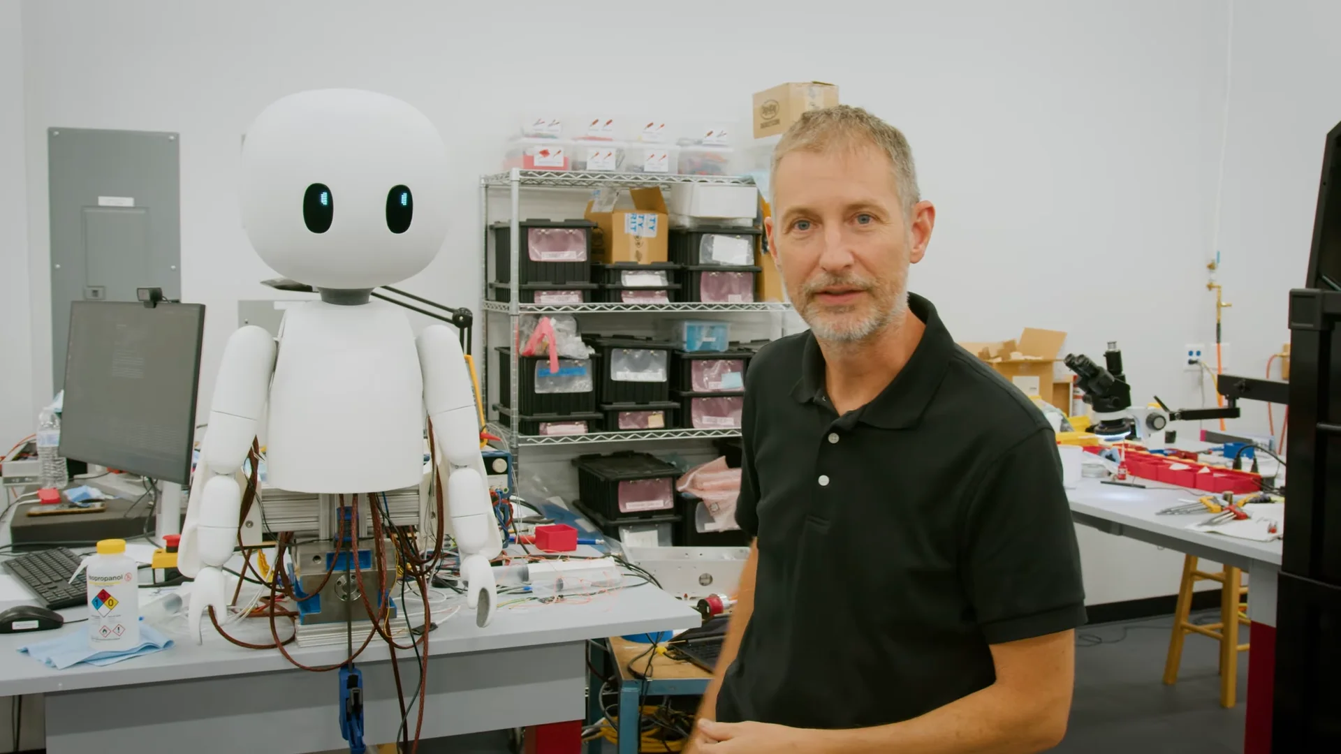 Cartwheel Robotics founder Scott LaValley stands inside a workshop next to the "Yogi" humanoid robot prototype. The robot features a large, round white head with illuminated blue eyes and an exposed torso revealing wiring and actuators.