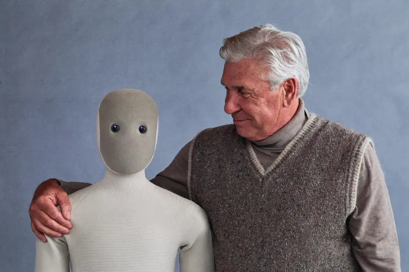 The 1X NEO humanoid robot stands side-by-side with an elderly man, who has his arm placed friendly on the robot's shoulder.