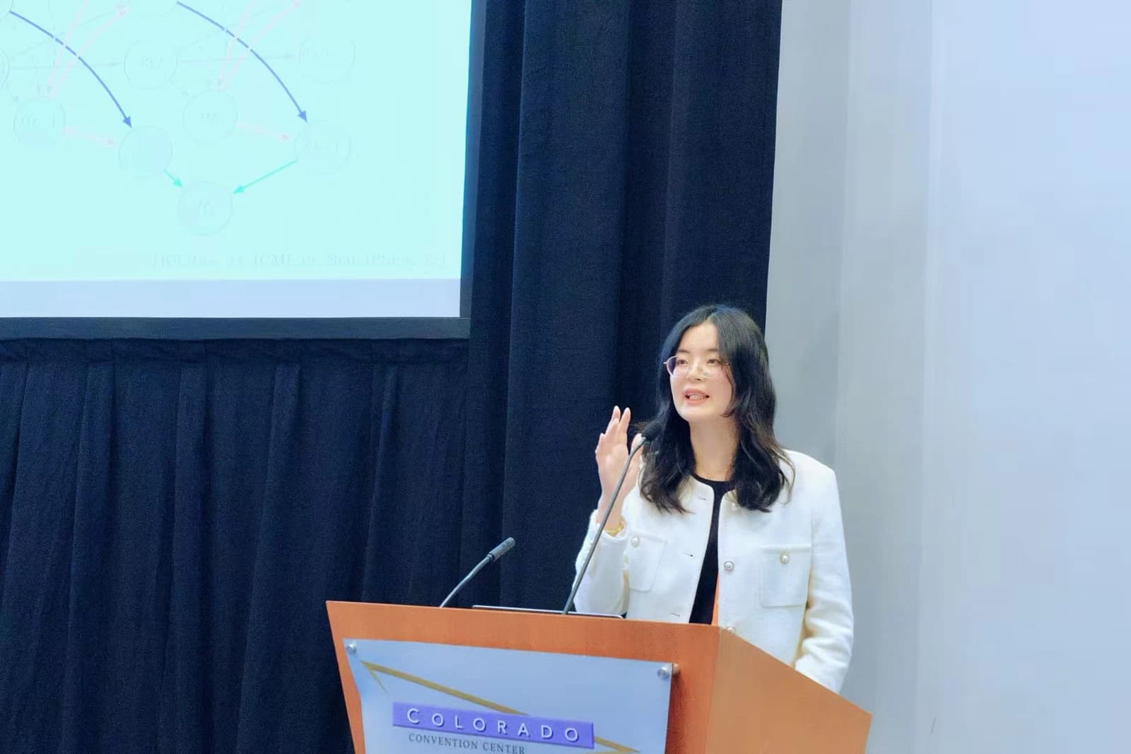 Professor Biwei Huang speaking at a lectern beside a presentation screen.
