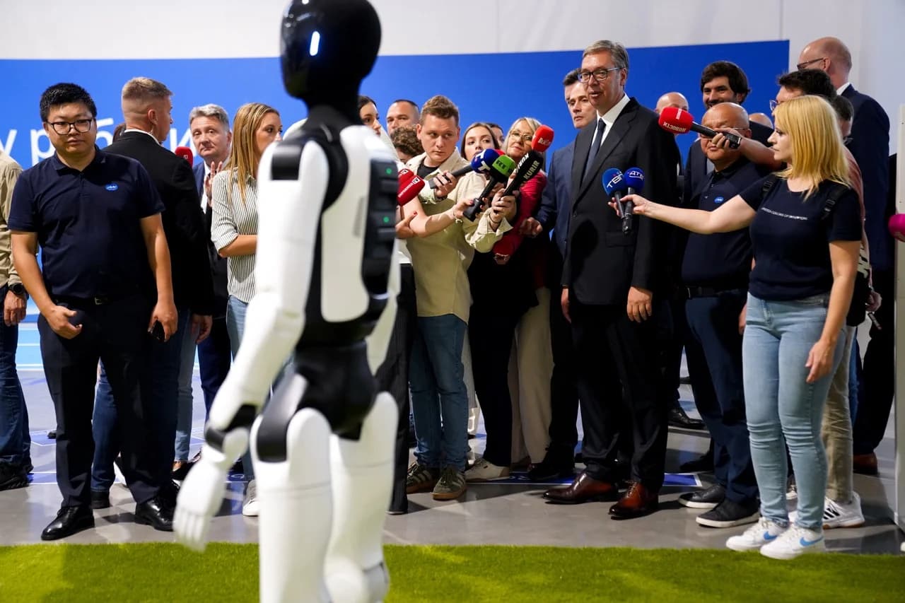 Serbian President Aleksandar Vučić speaks to a crowd of reporters holding microphones while an AGIBOT bipedal humanoid robot walks across artificial turf in the foreground during the launch event in Šabac.