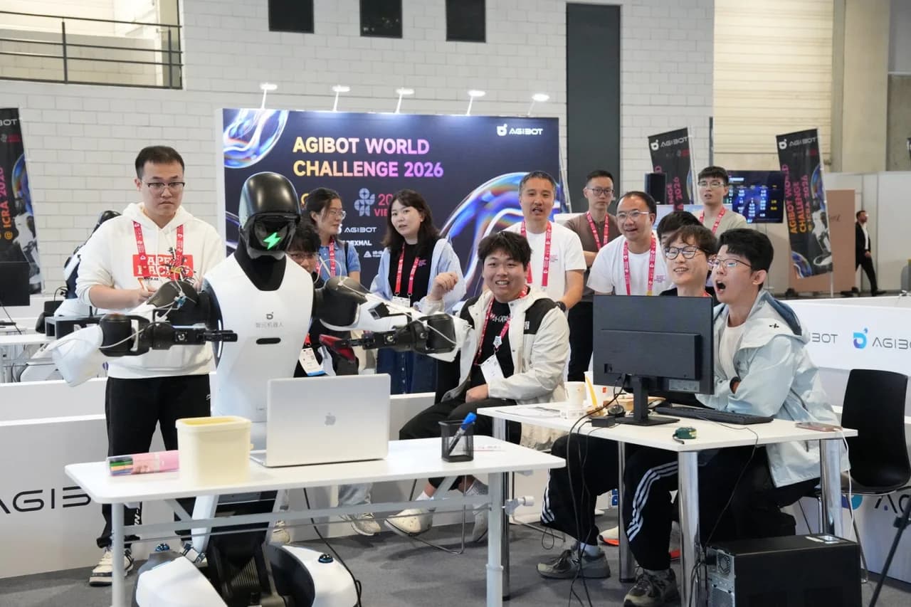 A team of participants sits at a desk cheering and smiling at a computer monitor next to an AGIBOT humanoid robot, with a crowd observing in the background.