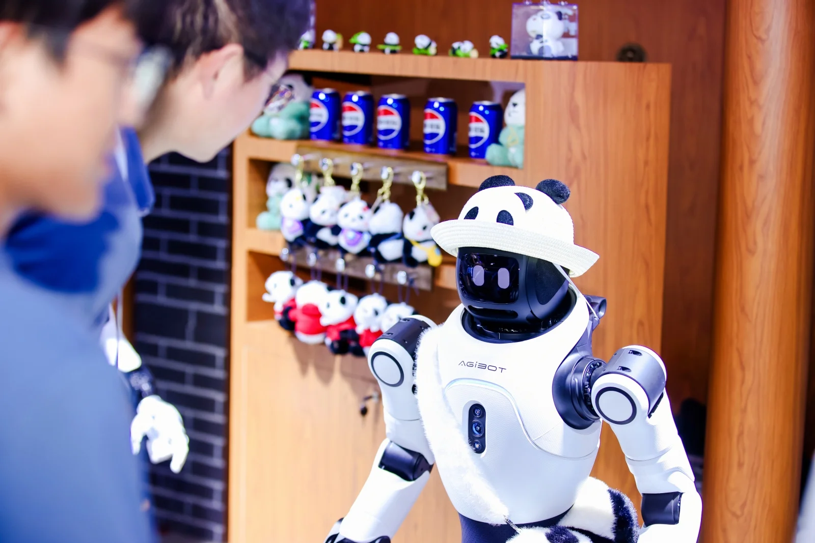A compact AGIBOT X2 EDU humanoid robot wearing a miniature panda hat and plush accessory while interacting with a visitor in front of merchandise shelves.