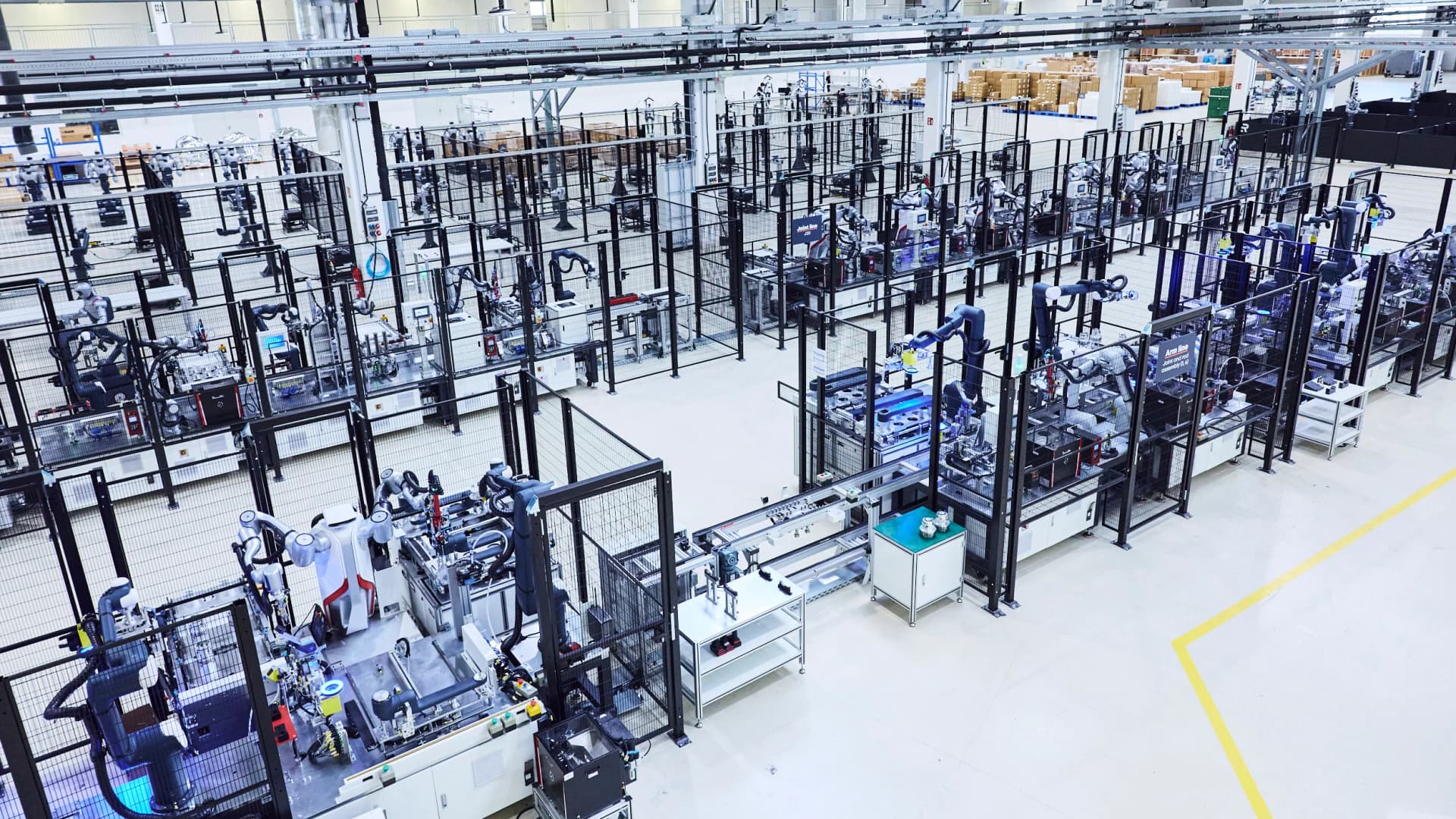 An elevated wide-angle view of a large, brightly lit industrial robotics facility with numerous safety-fenced assembly cells containing articulated robotic arms and mobile platforms.