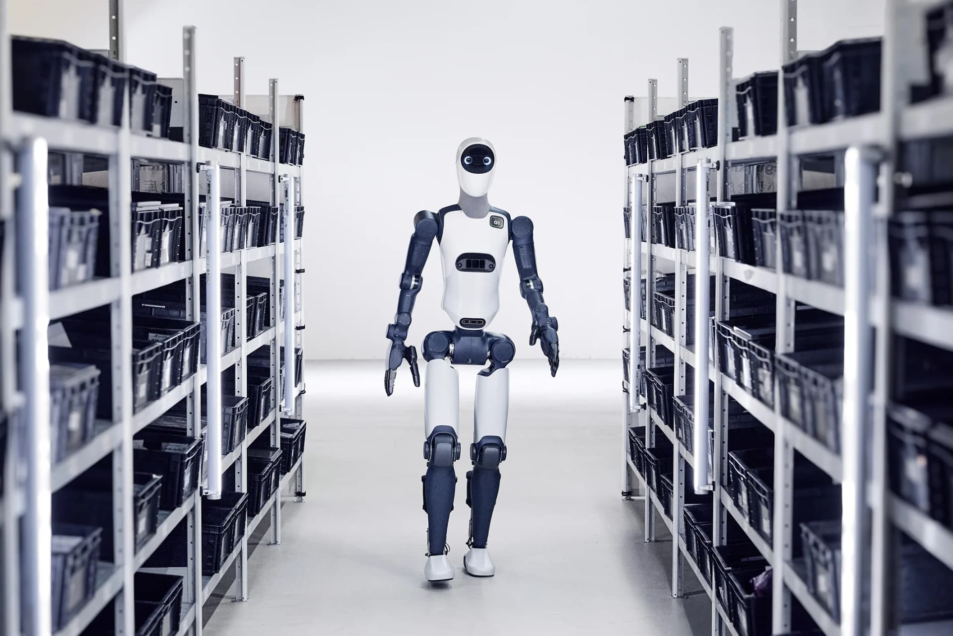 The bipedal Agile ONE humanoid robot walking down a narrow aisle between shelving units in a logistics facility.