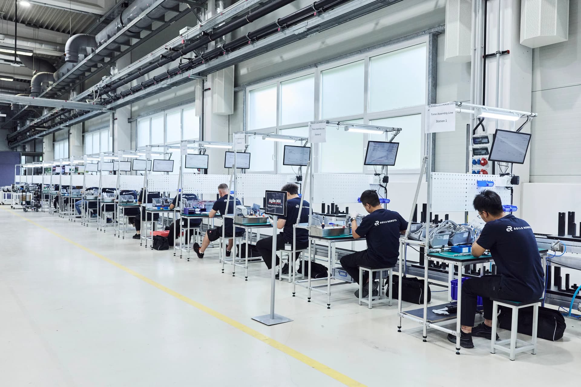 A row of technicians seated at white manual assembly benches labeled Torso Assembly Line along a factory floor, assembling subcomponents under monitor displays.