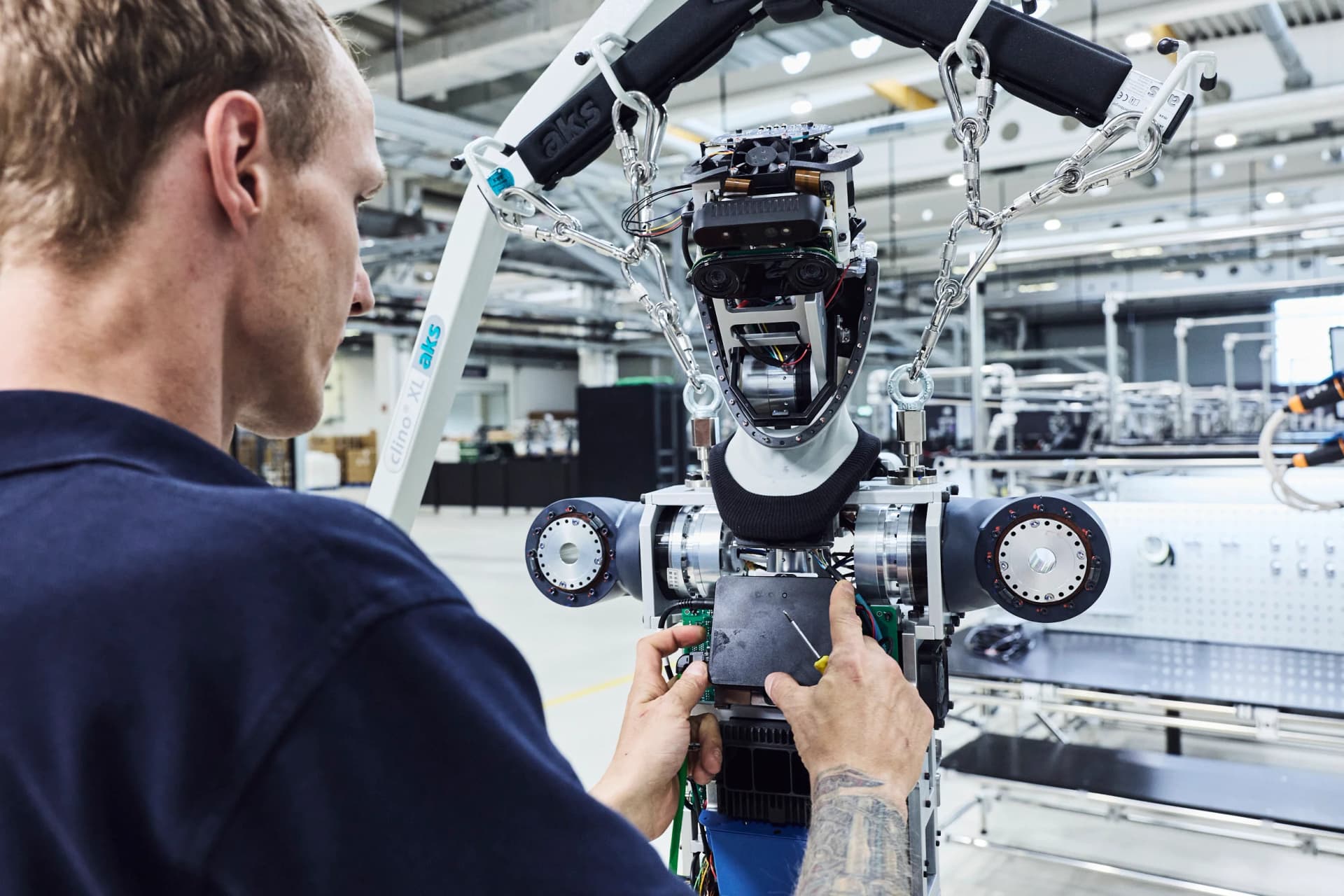 A technician in a navy blue shirt working on the internal electronics and torso wiring of an Agile ONE humanoid robot suspended from a white mechanical lift rig.