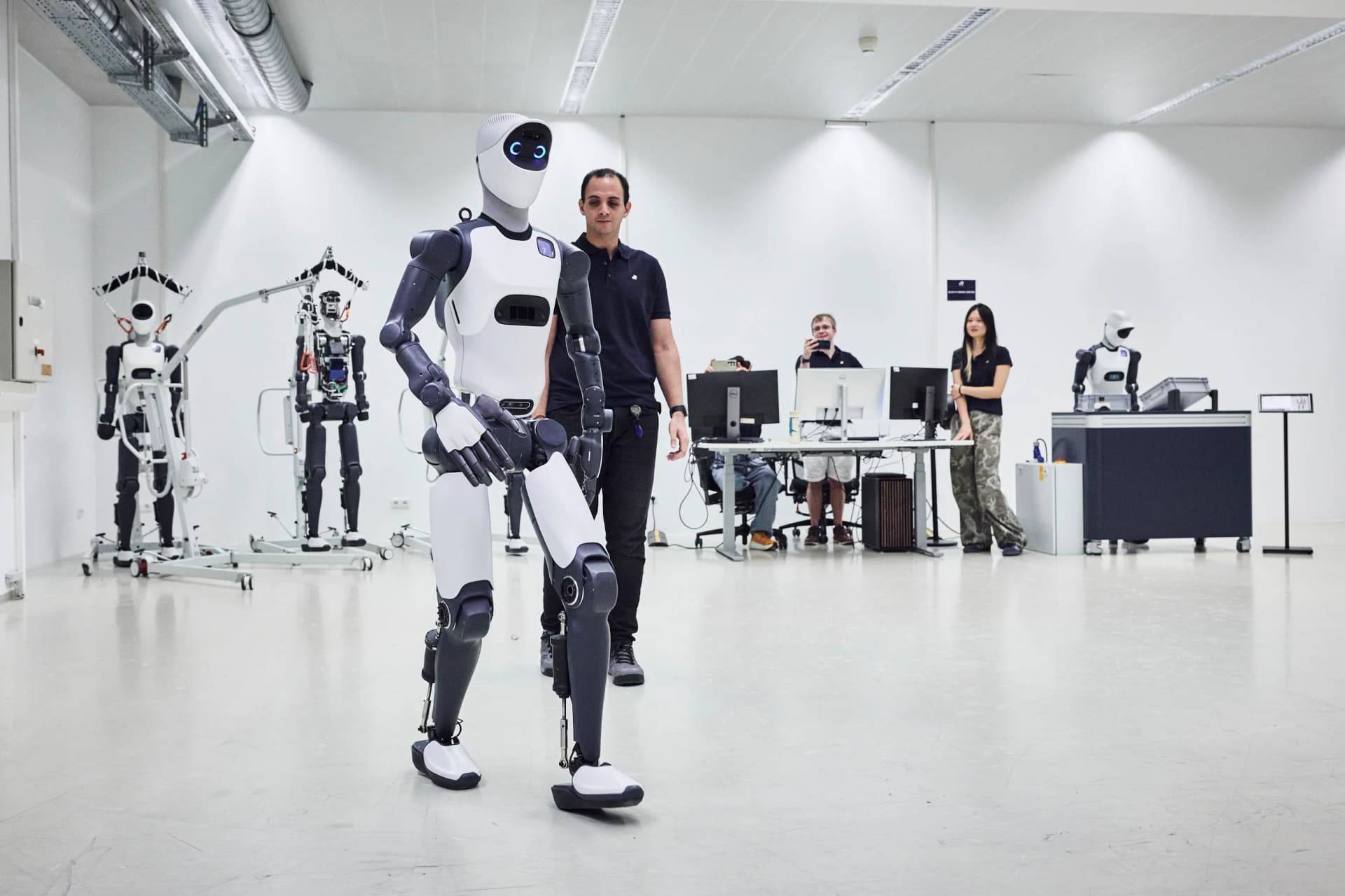The Agile ONE bipedal humanoid walking across a white testing laboratory floor, followed by an engineer while researchers observe from a workstation in the background.