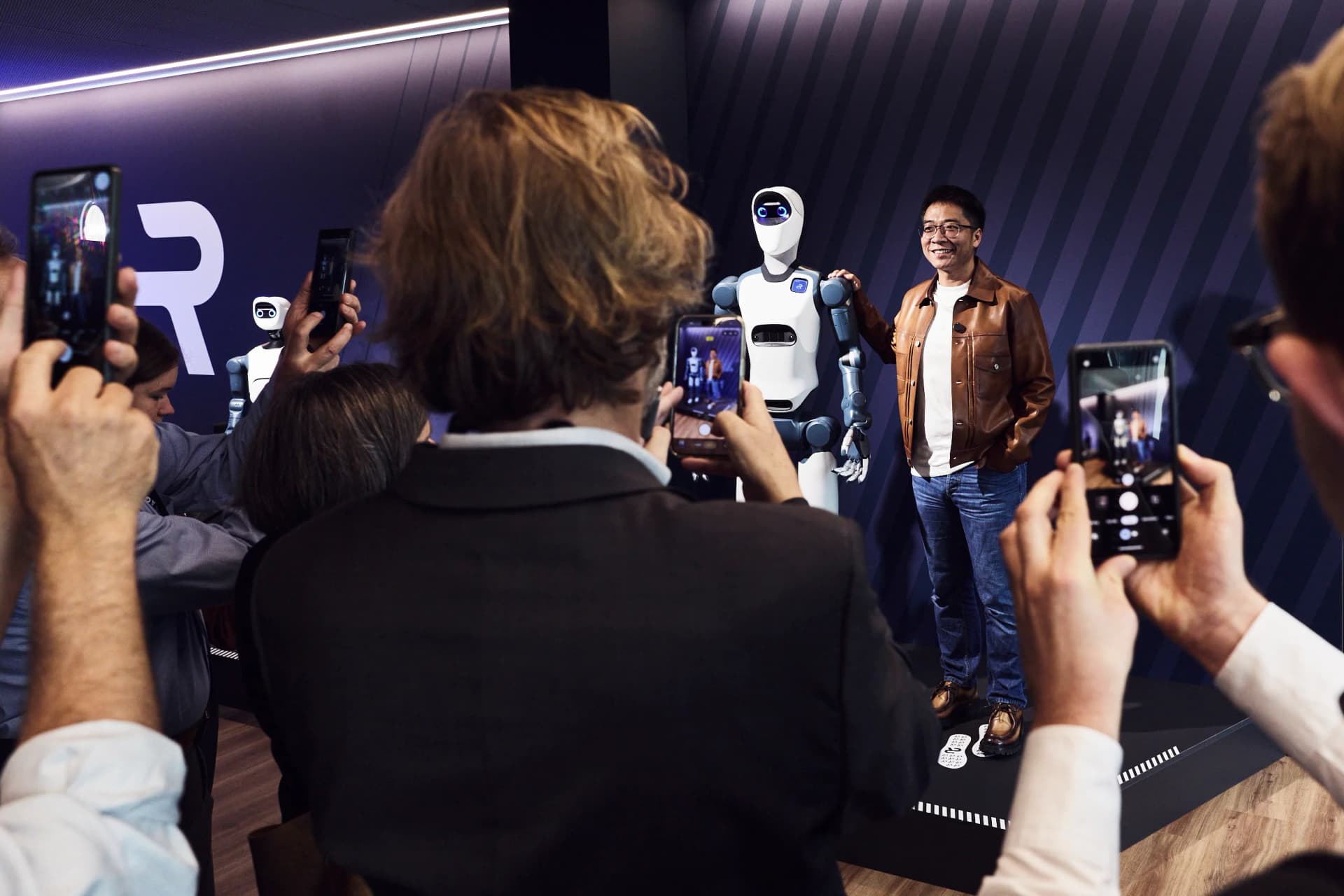 Agile Robots CEO Zhaopeng Chen wearing a brown leather jacket and standing next to the Agile ONE humanoid robot on a presentation stage while an audience photographs them.