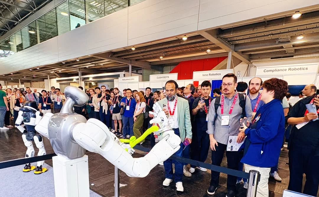 A dense crowd of attendees at an exhibition booth watching an AGILINK robotic arm manipulate a bright green balloon behind a safety barrier.