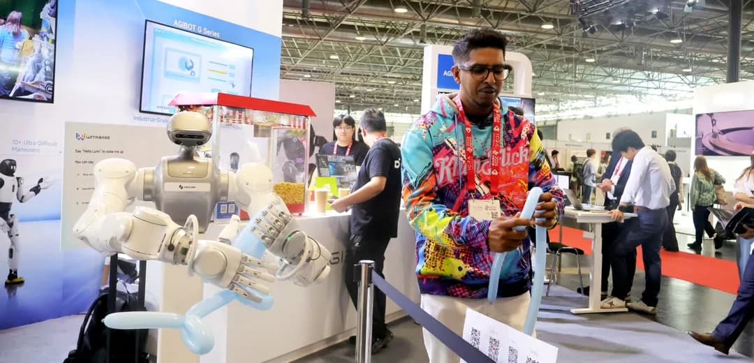 YouTuber KhanFlicks in a bright, multicolored hoodie stands next to an AGIBOT robot at a booth. Both are holding and manipulating light blue balloons side by side at ICRA 2026.