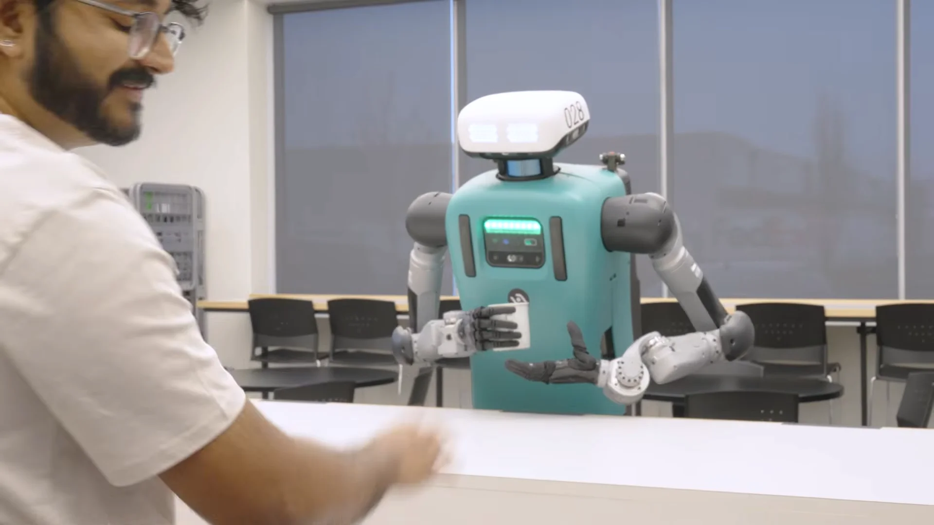 A still from an Agility promotional video showing the Digit humanoid robot (numbered 028) standing behind a long white counter in an office breakroom. On the left, a man with glasses and a beard looks toward the robot. Digit, with its familiar teal torso and white head, has been retrofitted with multi-fingered black robotic hands instead of its standard industrial end effectors. The robot holds a white coffee mug in its right hand while gesturing with its open left hand.