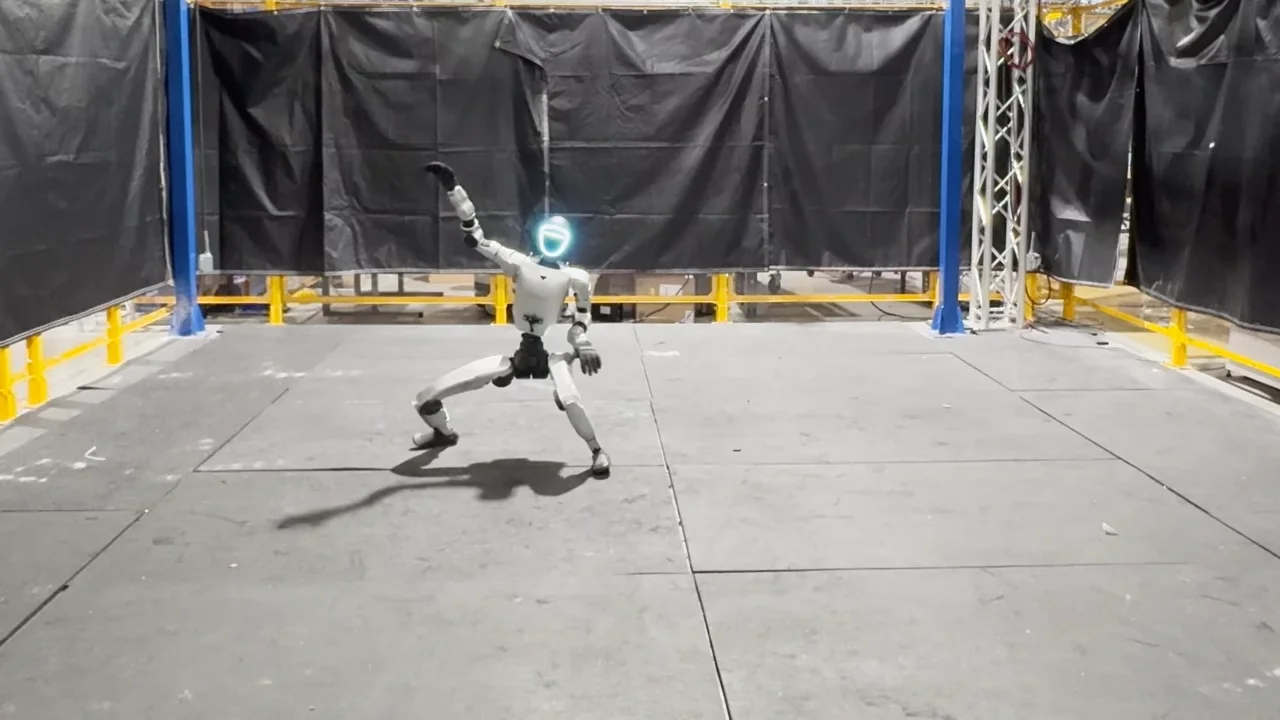 A Unitree G1 humanoid robot strikes a dynamic, wide-stance pose with one arm raised in a laboratory setting with yellow safety railings and black curtains, demonstrating a motion-tracking policy.