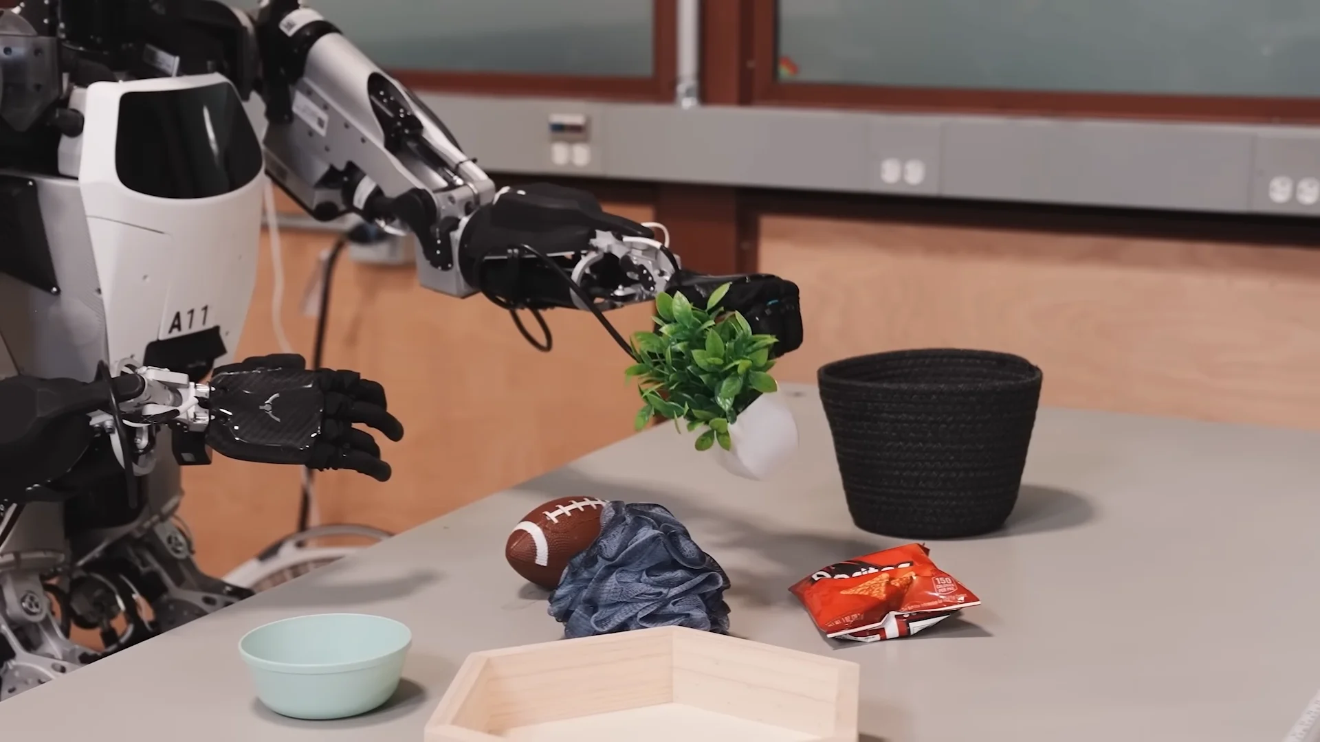 A robotic arm grasping a small potted plant from a table cluttered with various items like a football and a bag of chips.