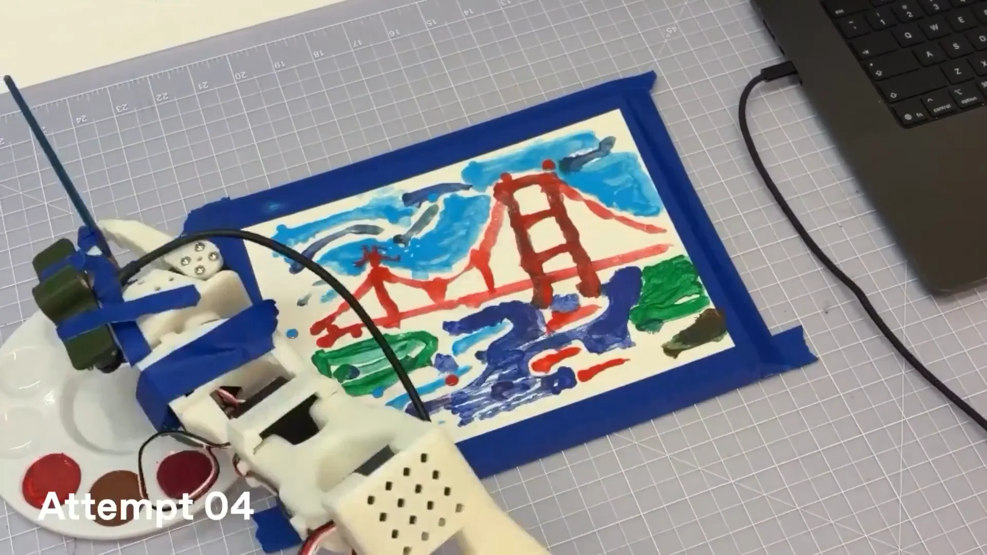 An overhead view of a 3D-printed robotic arm holding a thin blue paintbrush, painting a colorful depiction of the Golden Gate Bridge on paper taped to a cutting mat alongside a laptop and paint palette.
