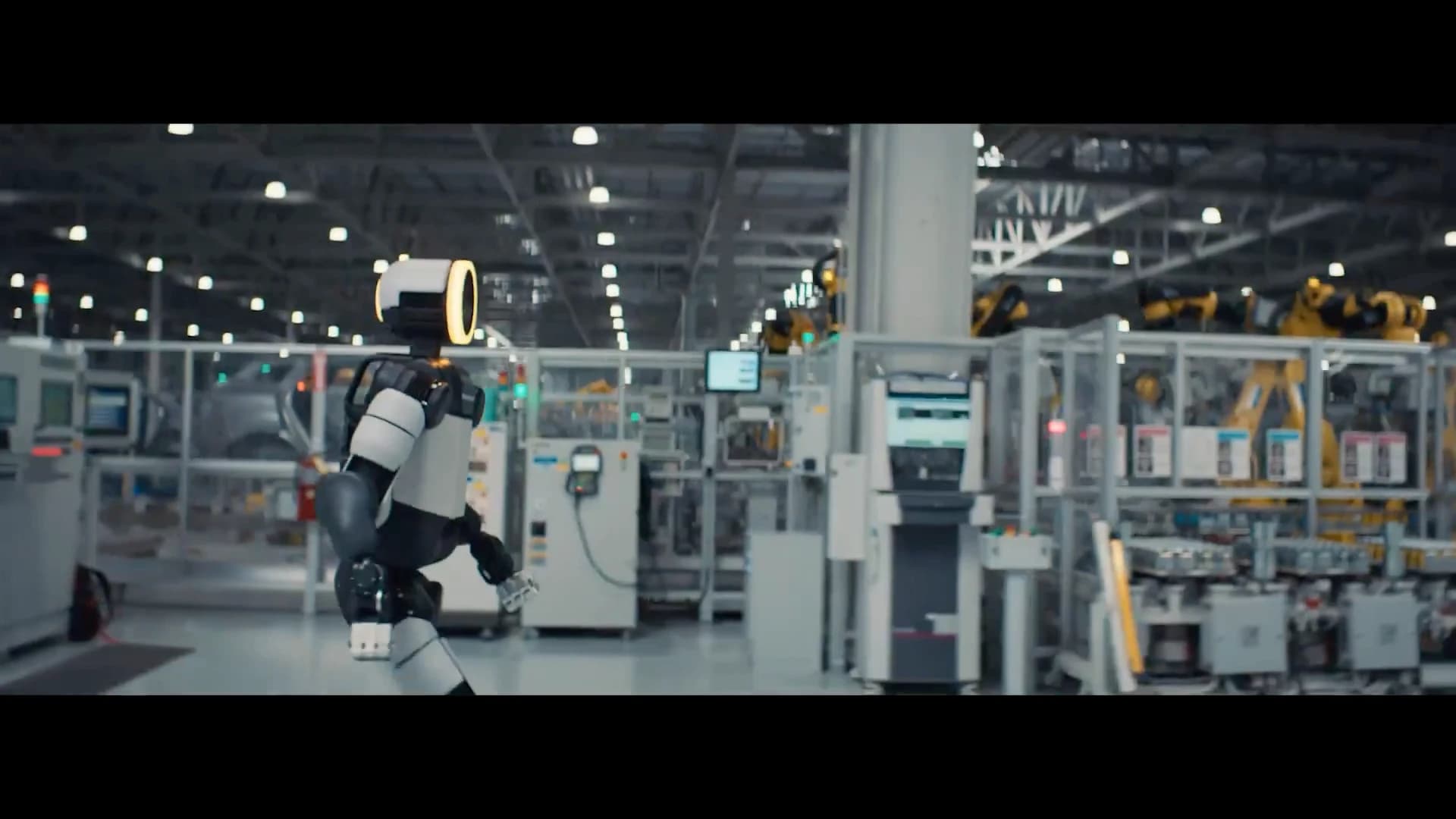 Atlas walks through a factory area with fenced industrial robot cells in the background.