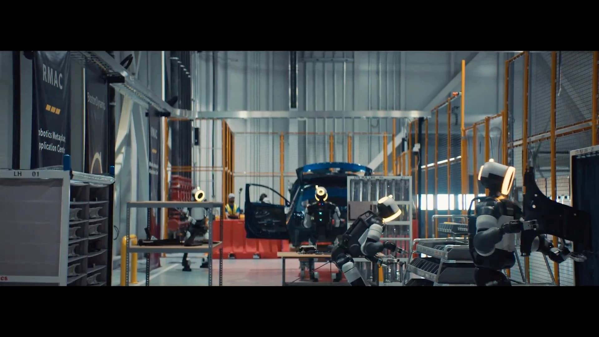 Several Atlas robots among parts racks and a vehicle inside the Robotics Metaplant Application Center.