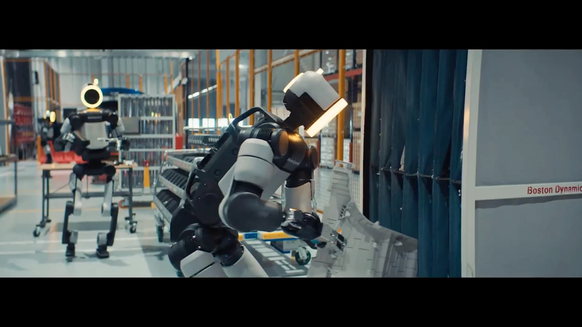 An Atlas robot handles a large molded component beside a storage rack, with other Atlas robots behind it.