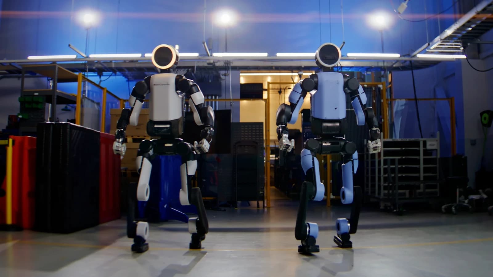 Two white-and-black Atlas humanoid robots standing side by side in a blue-lit robotics workspace.