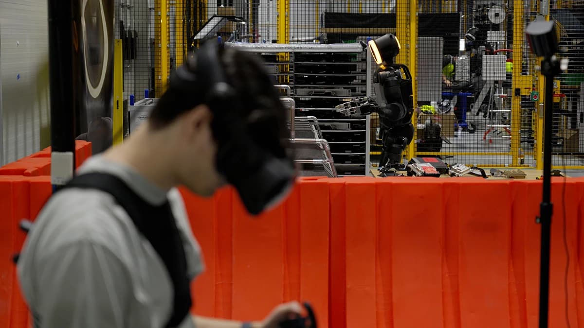 A trainer wearing a headset and holding a controller teleoperates Atlas at RMAC, with robots and parts racks in the background.