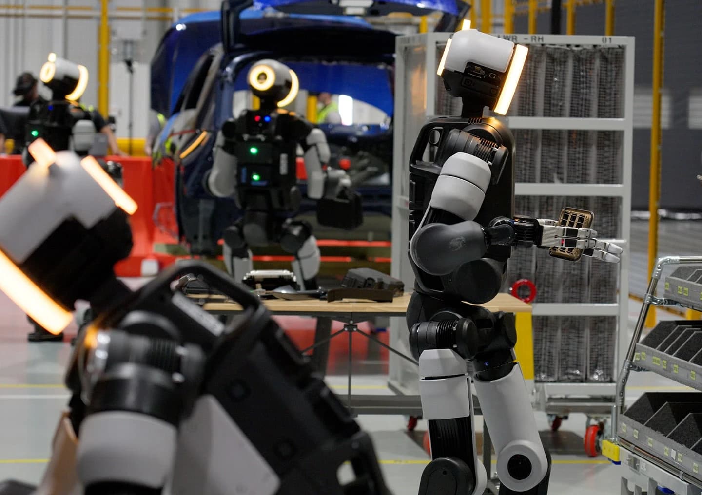Several Atlas robots practice handling parts among racks and workstations at the RMAC training center.