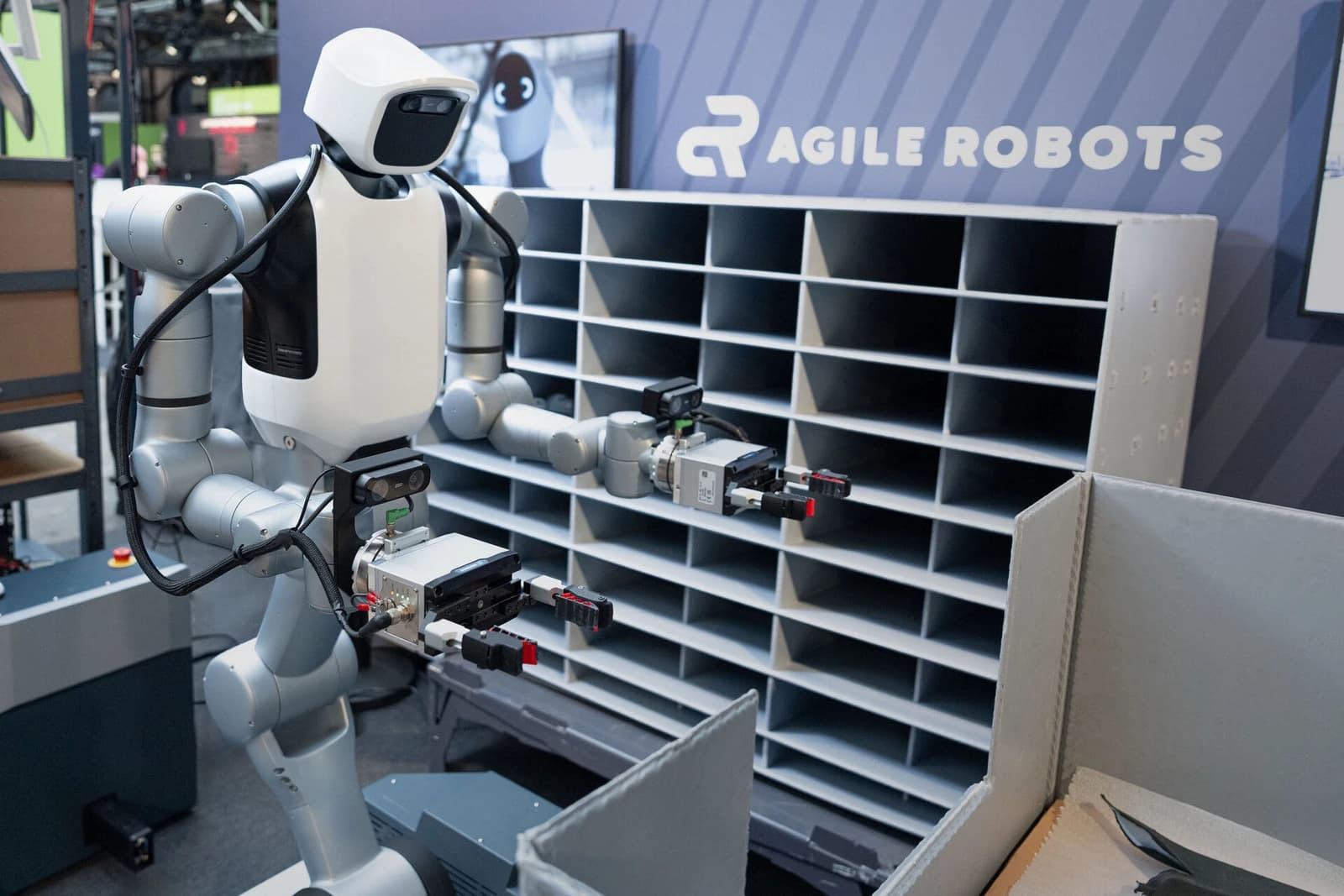 Agile Robots mobile humanoid with dual grippers beside parts containers and compartment shelving.